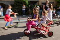 Zwei Mädchen die auf einem roten Dreirad sitzen. Im Hintergrund spielen weitere Kinder