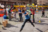 Spielende Kinder auf dem Marktplatz. Ein Junge spielt ein Zahlenspiel, das auf dem Boden gemalt ist. Hinter ihm promiert ein anderer Junge Steltzen aus.Stelzen