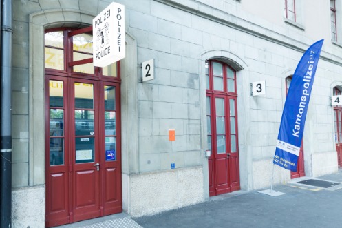 Vier hohe rote Türen aus Holz und Glas im hell gestalteten Eingangsbereich des Polizeiposten Bahnhof SBB lassen die Wache zum Hingucker werden. Ein blaues Banner und ein Schild über der Tür, weisen zusätzlich auf den Polizeiposten hin.
