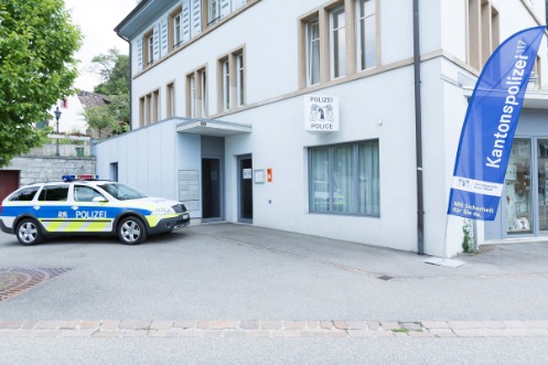 Dreistöckiges Gebäude des Polizeiposten Bettingen. Vor der Eingangstür parkt ein Polizeiwagen. Ein blaues Banner mit der Aufschrift "Kantonspolizei 117" und ein Schild mit dem Basler Wapppen und der Aufschrift "Polizei" zeichnen das weisse Gebäude als Polizeiwache aus.