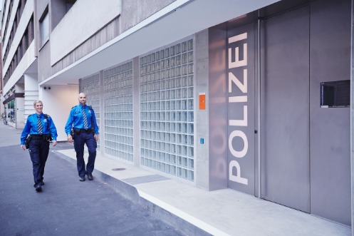 Eine Polizistin und ein Polizist auf dem Weg zum Eingang des städtischen Polizeiposten im Quartier Gundeldingen. Der moderne Eingangsbereich ist mit Glasbausteinen und einer Metaltür gestaltet, welche die Aufschrift "Polizei" ziert.