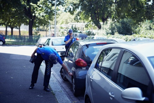 Zwei uniformierte Sicherheitsassistenten der Kantonspolizei Basel-Stadt bei der Kontrolle eines parkenden Kleinwagens auf einem städtischen Parkplatz neben einer Grünanlage.