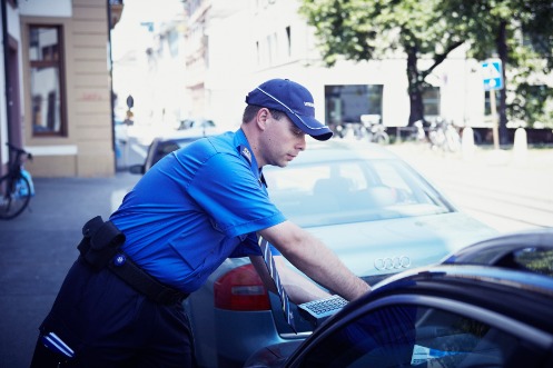 Sicherheitsassistent in Uniform und mit Capi bei der Prüfung der richtigen Parkausweisung in der Frontscheibe eines schwarzen Wagens.
